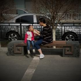 路边长椅上，一个母亲耐心的给女儿喂吃的，小女孩吃一...