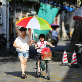 取景：小街上，小女孩在学骑自行车，妈妈撑着伞在边上...